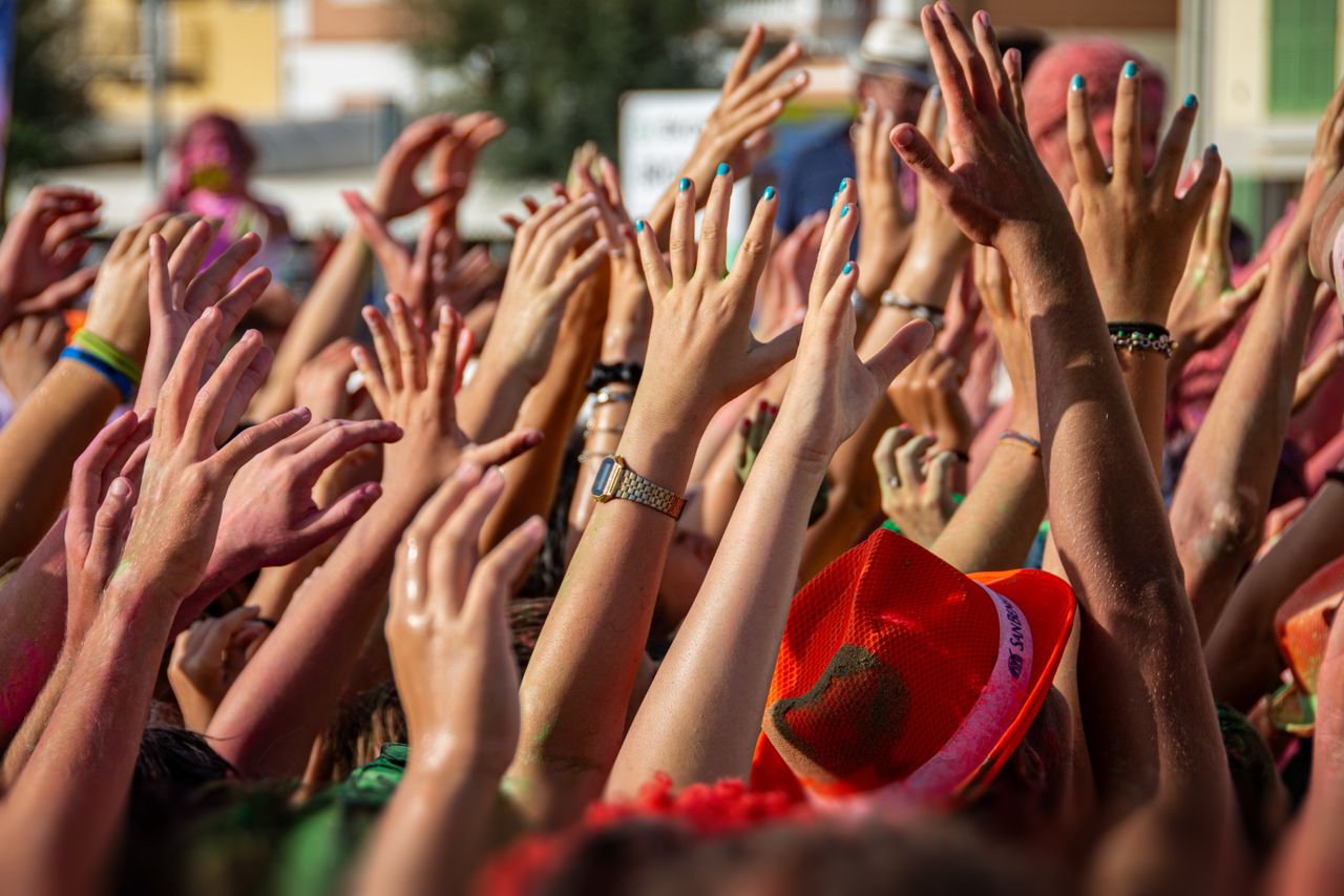 who-we-are A vibrant scene of people with colorful hands raised at an outdoor festival.